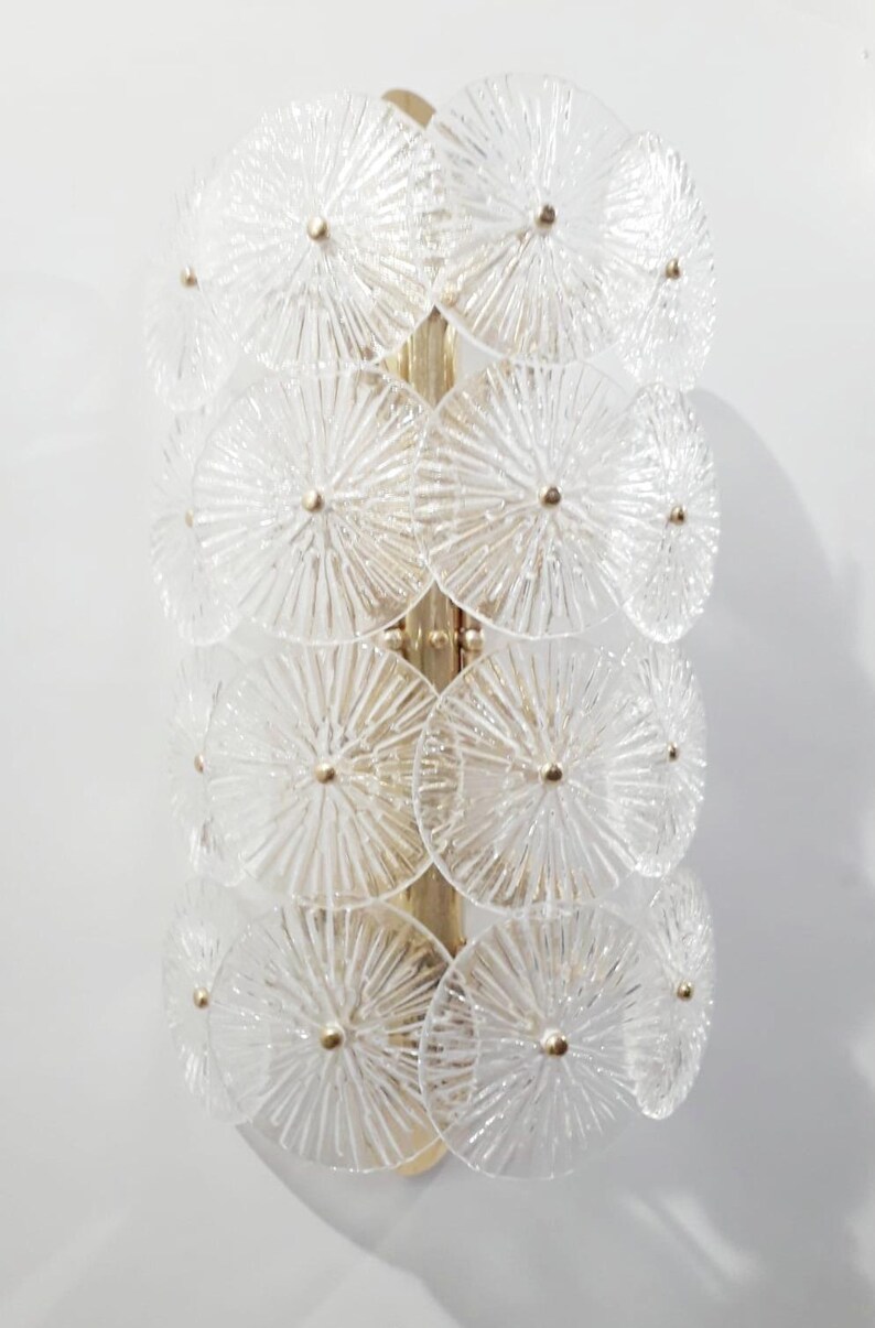 May include: A wall sconce featuring a vertical arrangement of clear, textured glass discs. The discs are arranged in a layered pattern and are attached to a gold-colored metal frame. Each disc has a small, gold-colored center.