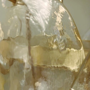 May include: Close-up of a clear glass chandelier with a textured surface. The glass has a yellowish tint and is attached to a gold-colored metal frame. The light fixture is likely for home decor.