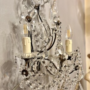 May include: Ornate wall sconce with clear crystal beads and floral accents. The sconce features two candle-style light bulbs and decorative crystal pendants. The metal frame has a dark finish, and the sconce is mounted on a wall.