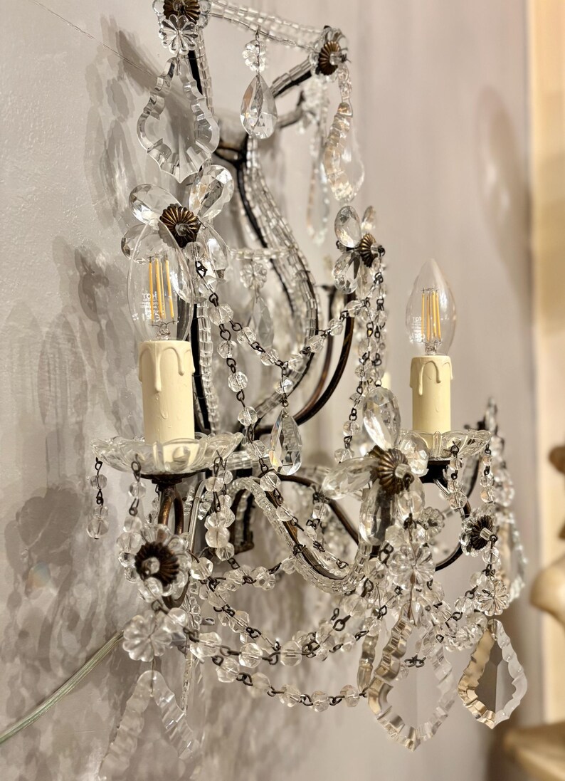 May include: Ornate wall sconce with clear crystal beads and floral accents. The sconce features two candle-style light bulbs and decorative crystal pendants. The metal frame has a dark finish, and the sconce is mounted on a wall.