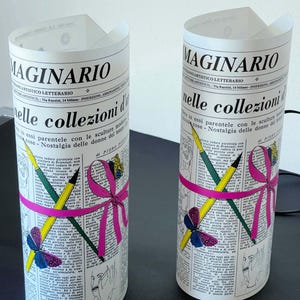 May include: Two white cylindrical lamps with a black and white newspaper print design. The lamps feature a pink ribbon with a bow, pencils, and a butterfly. The text on the lamps reads "Maginario" and "nelle collezioni d'arte".