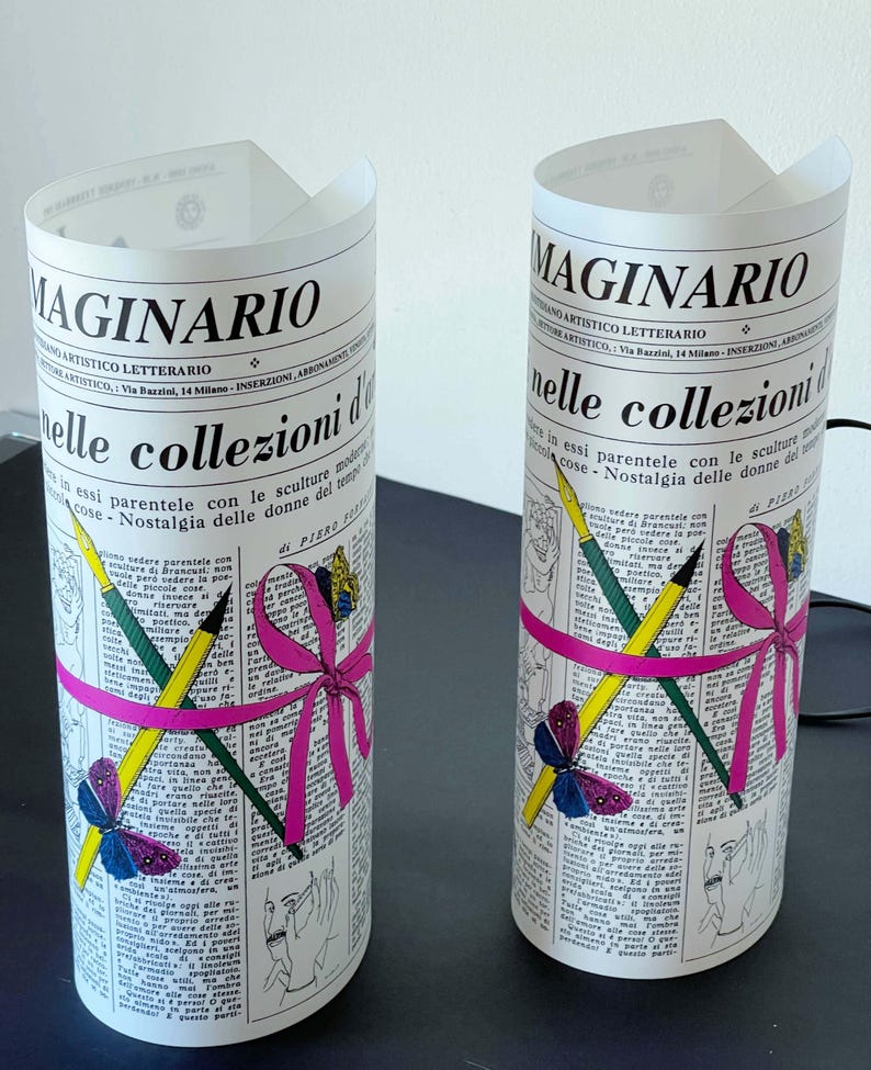 May include: Two white cylindrical lamps with a black and white newspaper print design. The lamps feature a pink ribbon with a bow, pencils, and a butterfly. The text on the lamps reads "Maginario" and "nelle collezioni d'arte".