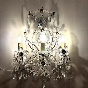 May include: A wall-mounted sconce with a vintage design. The sconce features clear crystal accents, including teardrop-shaped pendants and floral embellishments. Three light bulbs are visible, casting a warm glow. The metal frame has a dark finish.