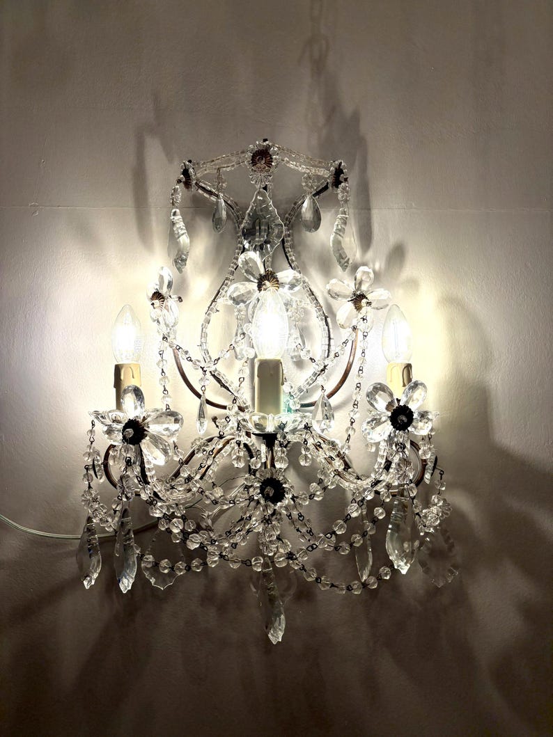 May include: A wall-mounted sconce with a vintage design. The sconce features clear crystal accents, including teardrop-shaped pendants and floral embellishments. Three light bulbs are visible, casting a warm glow. The metal frame has a dark finish.