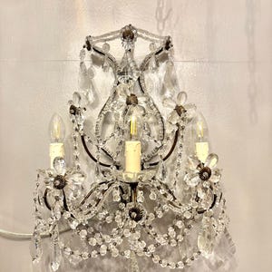 May include: An antique wall sconce with a dark metal frame, adorned with clear crystal beads and flower-shaped accents. The sconce features three light bulbs and hanging crystal pendants, creating a vintage aesthetic.