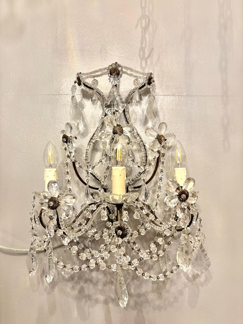 May include: An antique wall sconce with a dark metal frame, adorned with clear crystal beads and flower-shaped accents. The sconce features three light bulbs and hanging crystal pendants, creating a vintage aesthetic.
