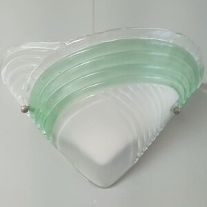 May include: A white and green glass wall sconce with a geometric design. The sconce has a triangular shape and features a green glass band with a wavy pattern.