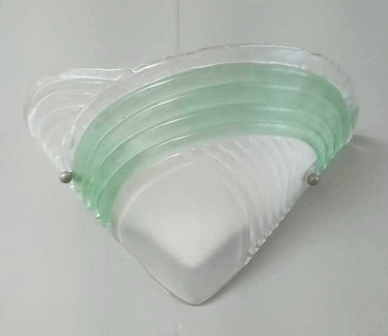 May include: A white and green glass wall sconce with a geometric design. The sconce has a triangular shape and features a green glass band with a wavy pattern.