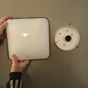 May include: A white square ceiling light fixture with a silver trim. The light fixture is attached to a white ceiling with a silver mounting plate.