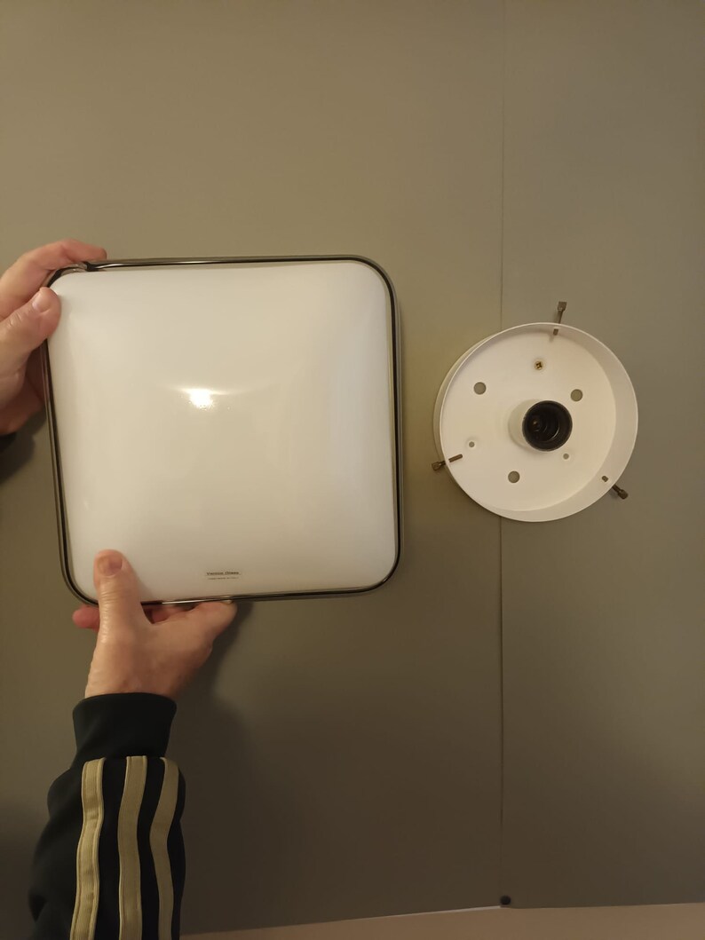 May include: A white square ceiling light fixture with a silver trim. The light fixture is attached to a white ceiling with a silver mounting plate.