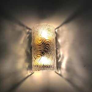 May include: A clear glass wall sconce with a gold swirl pattern. The sconce has two light bulbs and is mounted on a white wall.