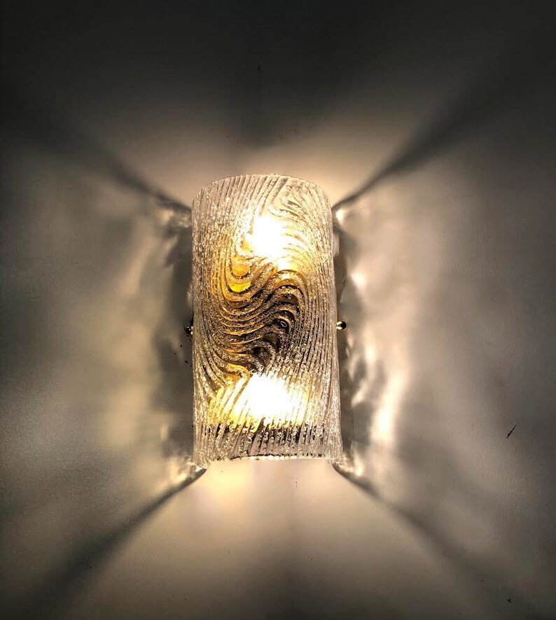 May include: A clear glass wall sconce with a gold swirl pattern. The sconce has two light bulbs and is mounted on a white wall.