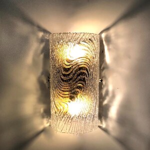 May include: A frosted glass wall sconce with a wavy pattern and two light bulbs. The sconce is mounted on a white wall.