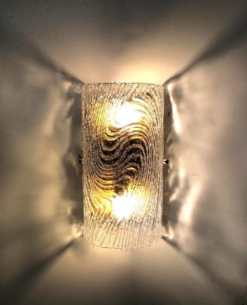 May include: A frosted glass wall sconce with a wavy pattern and two light bulbs. The sconce is mounted on a white wall.