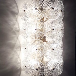 May include: A wall-mounted light fixture with a series of clear, circular glass elements. Each disc has a textured, radial pattern and a dark center. The fixture is mounted on a gold-toned metal frame, casting light and shadows.