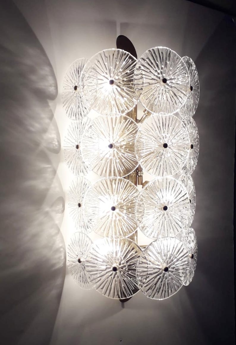 May include: A wall-mounted light fixture with a series of clear, circular glass elements. Each disc has a textured, radial pattern and a dark center. The fixture is mounted on a gold-toned metal frame, casting light and shadows.
