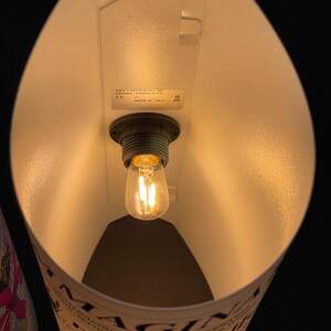 May include: A white lampshade with a light bulb inside. The lampshade is made of paper and has a printed design. The text on the lampshade reads "MAGNIFICHE" and "delle citt&agrave;".