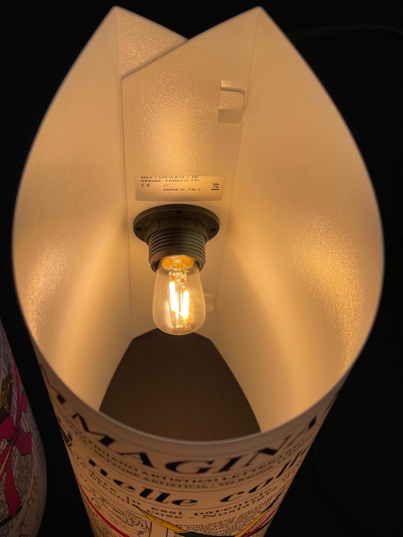 May include: A white lampshade with a light bulb inside. The lampshade is made of paper and has a printed design. The text on the lampshade reads "MAGNIFICHE" and "delle citt&agrave;".