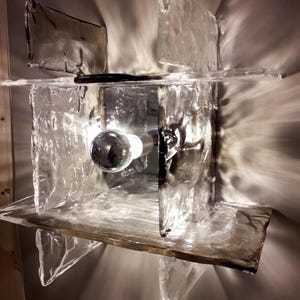 May include: A modern wall sconce with a geometric design made of clear glass panels. The sconce has a single light bulb in the centre.