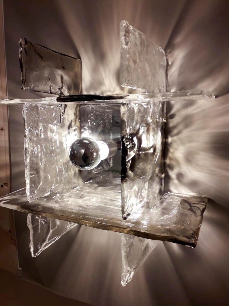 May include: A modern wall sconce with a geometric design made of clear glass panels. The sconce has a single light bulb in the centre.