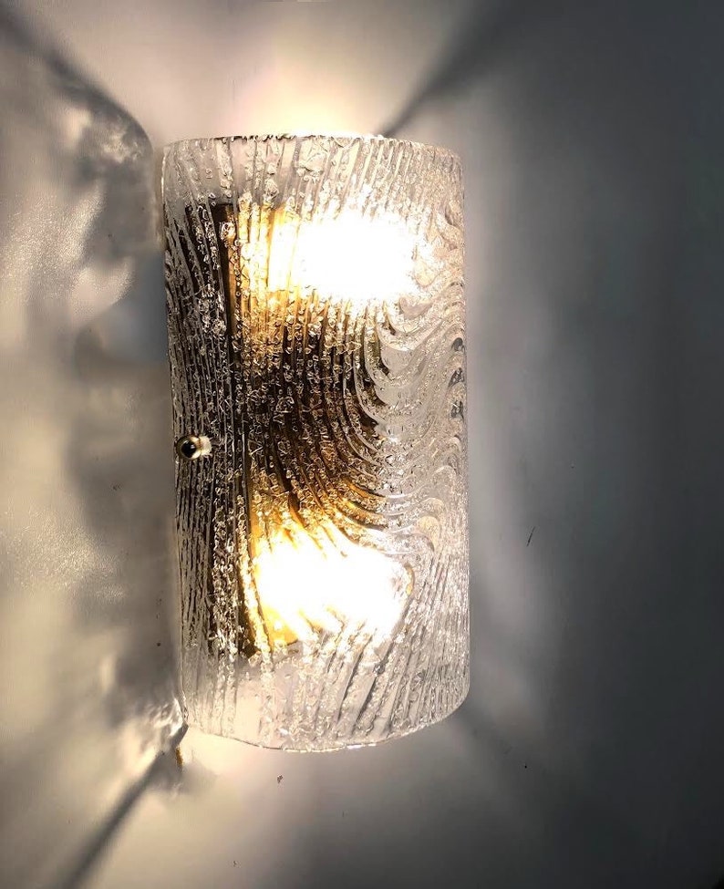 May include: A clear glass wall sconce with a textured, wavy pattern. The sconce has a brass backing and two light bulbs.