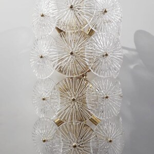 May include: A vertical wall sconce featuring a series of clear, textured glass discs arranged in a layered pattern. The discs are connected by gold-toned metal supports, creating a decorative lighting fixture. The glass has a radial design.