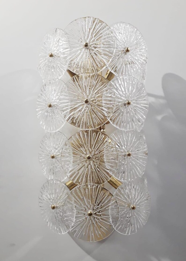 May include: A vertical wall sconce featuring a series of clear, textured glass discs arranged in a layered pattern. The discs are connected by gold-toned metal supports, creating a decorative lighting fixture. The glass has a radial design.