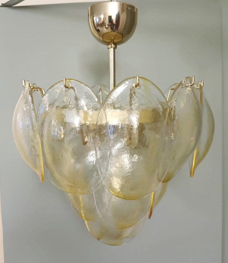May include: A vintage-style chandelier with a gold-toned metal frame and multiple tiers of textured, petal-shaped glass shades. The glass has a soft yellow hue, creating a warm, inviting glow. The fixture is suspended from a matching gold ceiling mount.