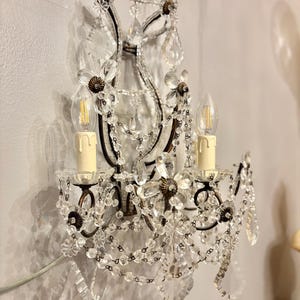 May include: An ornate wall sconce with two candle-style light bulbs. The sconce features a dark metal frame adorned with clear crystal beads, flower-shaped accents, and hanging crystal pendants. The sconce is mounted on a light-colored wall.