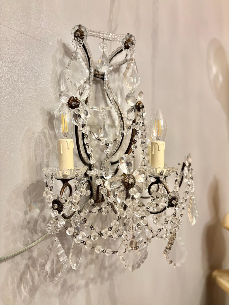 May include: An ornate wall sconce with two candle-style light bulbs. The sconce features a dark metal frame adorned with clear crystal beads, flower-shaped accents, and hanging crystal pendants. The sconce is mounted on a light-colored wall.