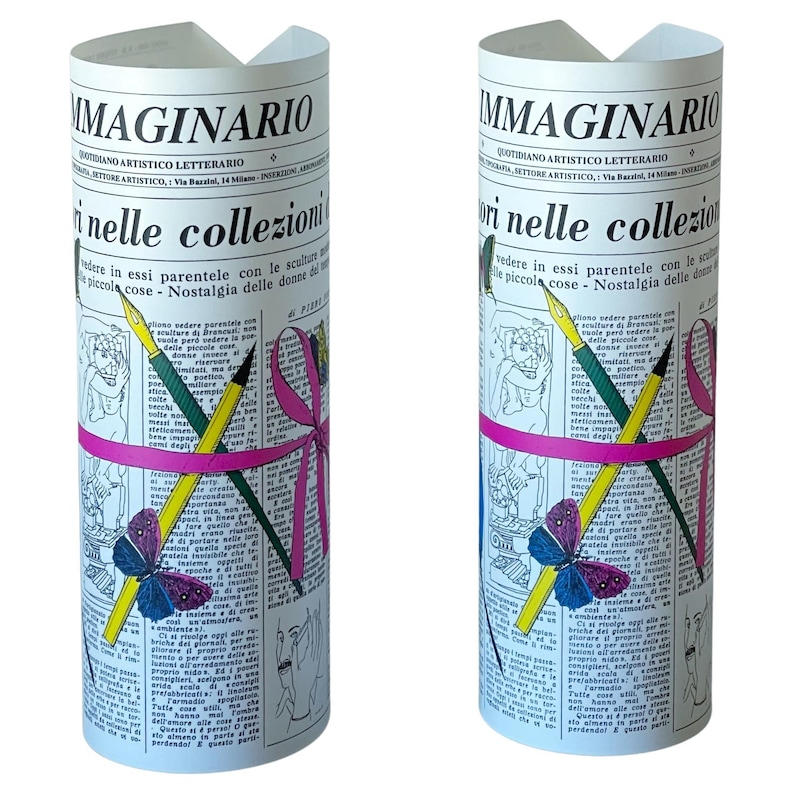 May include: Two white cylindrical candles with a newspaper design. The candles feature a pink ribbon, a butterfly, and pencils. The text on the candles reads "IMMAGINARIO" and "QUOTIDIANO ARTISTICO LETTERARIO".