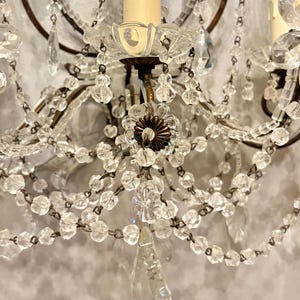 May include: Close-up of a vintage wall sconce with clear crystal beads and teardrop pendants. The sconce features a brown metal frame, candle-shaped light fixtures, and a decorative floral element. The design is ornate and elegant.