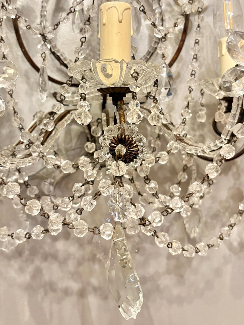 May include: Close-up of a vintage wall sconce with clear crystal beads and teardrop pendants. The sconce features a brown metal frame, candle-shaped light fixtures, and a decorative floral element. The design is ornate and elegant.