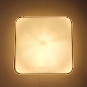 May include: A white square ceiling light fixture with a rounded edge. The light is on and emits a warm, yellow glow. The text "Venini Glass Made in Italy" is printed on the bottom of the fixture.