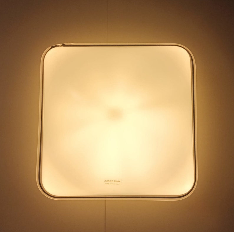 May include: A white square ceiling light fixture with a rounded edge. The light is on and emits a warm, yellow glow. The text "Venini Glass Made in Italy" is printed on the bottom of the fixture.