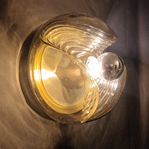 Puede incluir: Una l&aacute;mpara de pared de vidrio de color dorado con un dise&ntilde;o redondo y ondulado. La l&aacute;mpara tiene una sola bombilla en el centro.