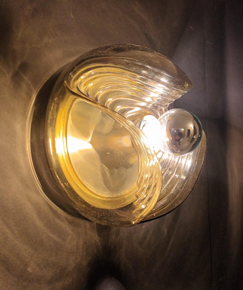 Puede incluir: Una l&aacute;mpara de pared de vidrio de color dorado con un dise&ntilde;o redondo y ondulado. La l&aacute;mpara tiene una sola bombilla en el centro.