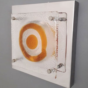 May include: A square wall sconce with a frosted glass panel featuring a concentric circle design in shades of amber and white. The sconce is mounted on a silver metal frame.