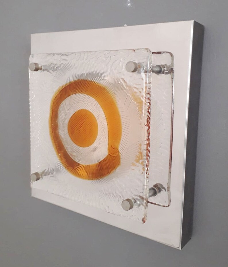 May include: A square wall sconce with a frosted glass panel featuring a concentric circle design in shades of amber and white. The sconce is mounted on a silver metal frame.