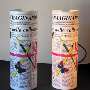 May include: Two white cylindrical lamps with a newspaper design. The lamps feature a pink ribbon, a butterfly, and pencils and pens. The text on the lamps reads "IMMAGINARIO" and "QUOTIDIANO ARTISTICO LETTERARIO".