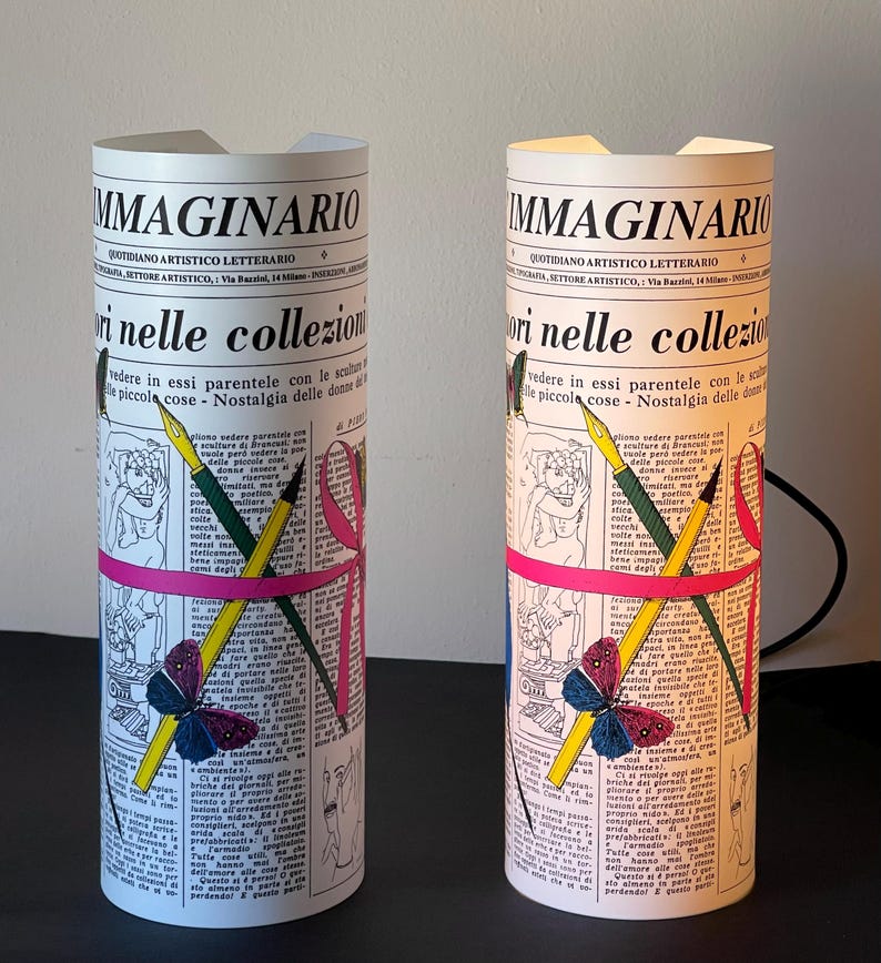 May include: Two white cylindrical lamps with a newspaper design. The lamps feature a pink ribbon, a butterfly, and pencils and pens. The text on the lamps reads "IMMAGINARIO" and "QUOTIDIANO ARTISTICO LETTERARIO".