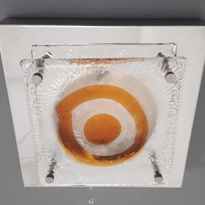 May include: A square ceiling light fixture with a chrome frame and a frosted glass shade. The glass shade has a concentric circle design with an orange center.