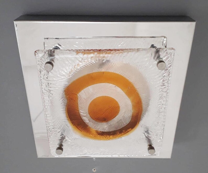 May include: A square ceiling light fixture with a chrome frame and a frosted glass shade. The glass shade has a concentric circle design with an orange center.