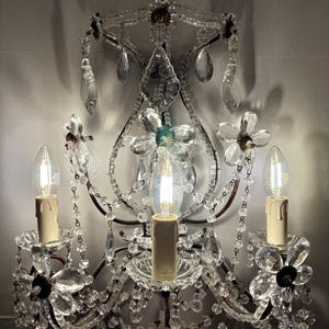 May include: An ornate wall sconce with three candle-style light bulbs. The sconce features clear crystal beads, flower-shaped accents, and hanging crystal droplets. The metal frame has a dark, antique finish, and the bulbs are lit.