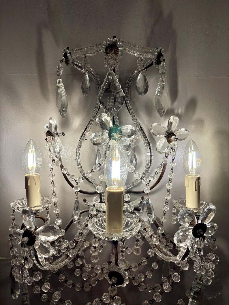 May include: An ornate wall sconce with three candle-style light bulbs. The sconce features clear crystal beads, flower-shaped accents, and hanging crystal droplets. The metal frame has a dark, antique finish, and the bulbs are lit.