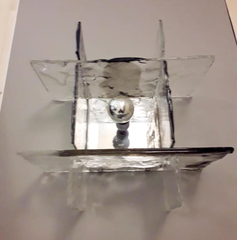 May include: A modern glass and metal ceiling light fixture with a geometric design. The light fixture has a clear glass shade with a silver metal base.
