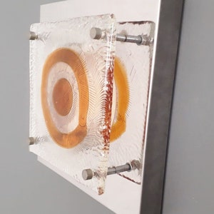 May include: A wall-mounted light fixture with a square, frosted glass shade. The shade features a concentric circle design in shades of orange and white.