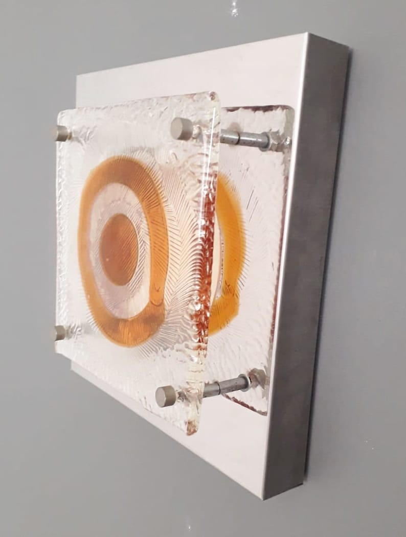 May include: A wall-mounted light fixture with a square, frosted glass shade. The shade features a concentric circle design in shades of orange and white.