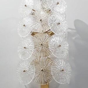 May include: A wall sconce featuring a series of clear, circular glass elements arranged in a vertical pattern. The glass discs have a textured, radial design and are attached to a gold-toned metal frame. The sconce is designed to be mounted on a wall.