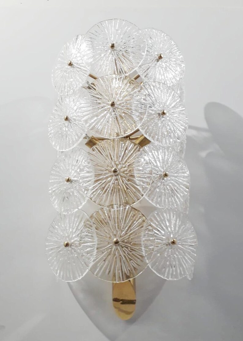 May include: A wall sconce featuring a series of clear, circular glass elements arranged in a vertical pattern. The glass discs have a textured, radial design and are attached to a gold-toned metal frame. The sconce is designed to be mounted on a wall.